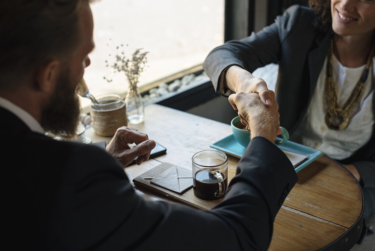 Business handshake across a table — rawpixel.com, CC0, via Wikimedia Commons